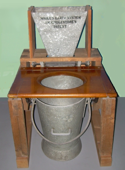 Large bucket under wooden seat with hole, metal hopper above and behind